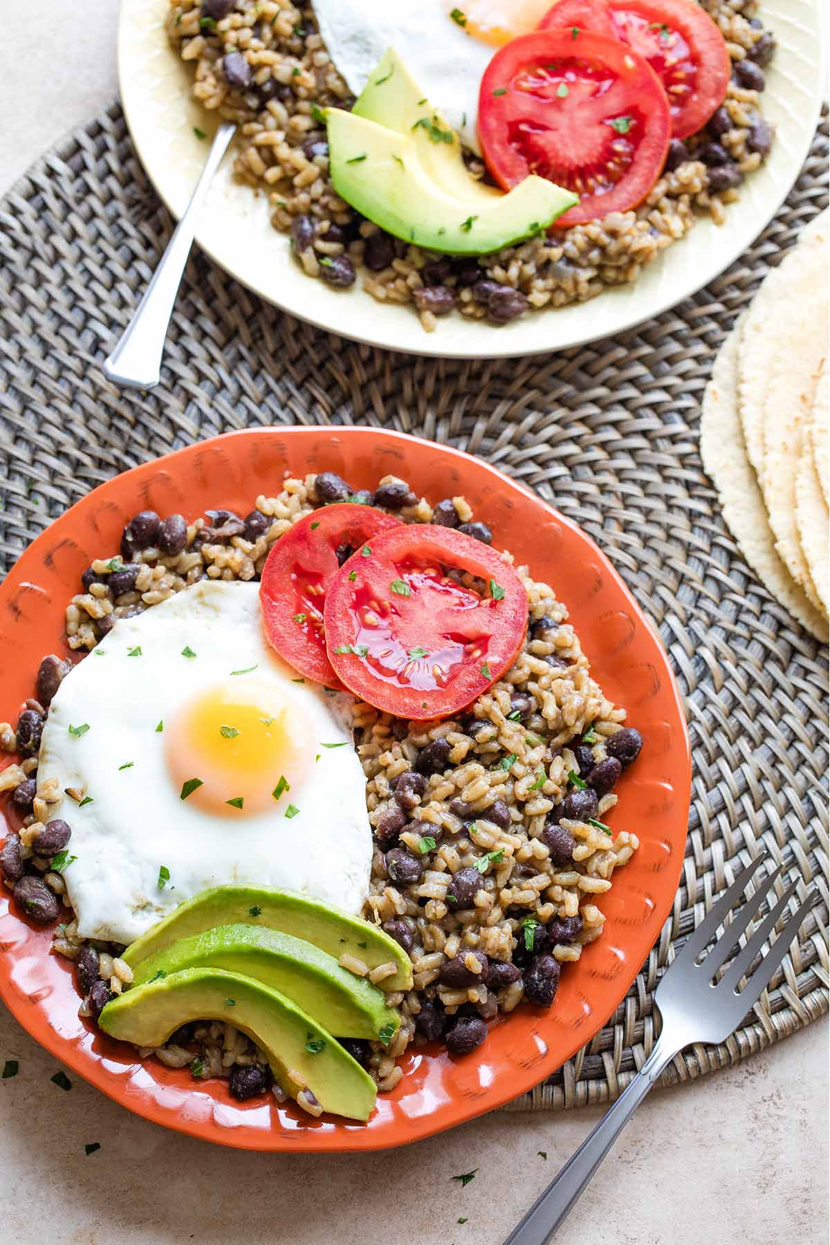Flatlay of two platefuls of Gallo Pinto with toppings, sitting on round woven mat with forks and stack of tortillas nearby.