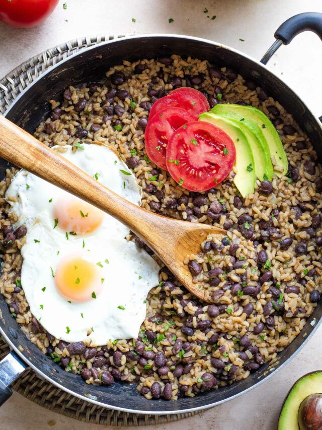 Overhead of cooked Gallo Pinto in pan with minced cilantro, avocado and tomato slices, 2 fried eggs on top.