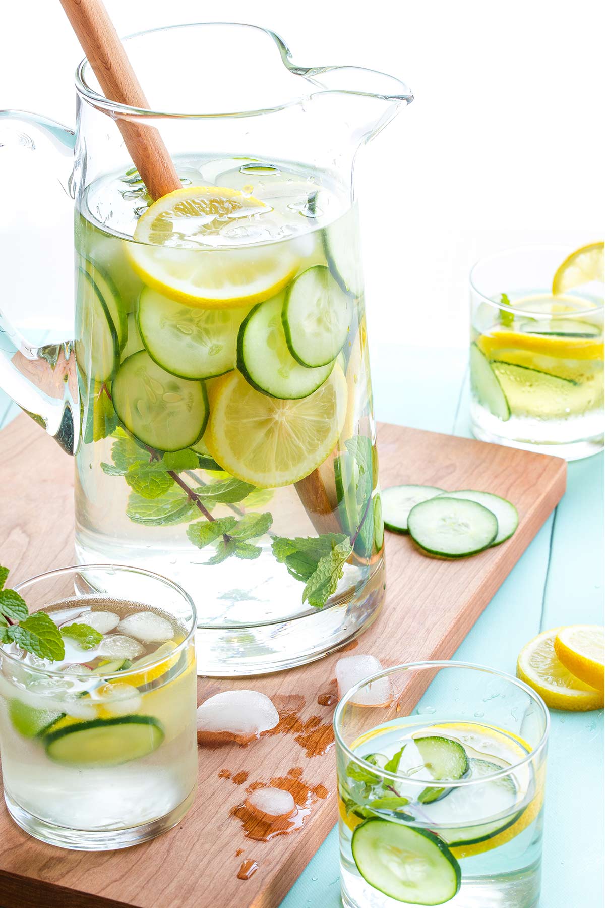 Pitcher surrounded by 3 glasses of water, with melting ice and extra cucumber and lemon slices scattered around.