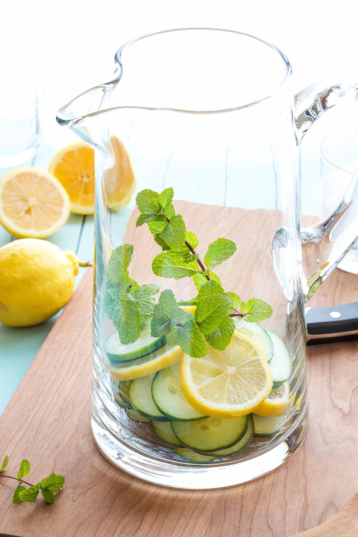 Slices of cucumber and lemon piled in glass pitcher with a couple mint sprigs, before water is added.