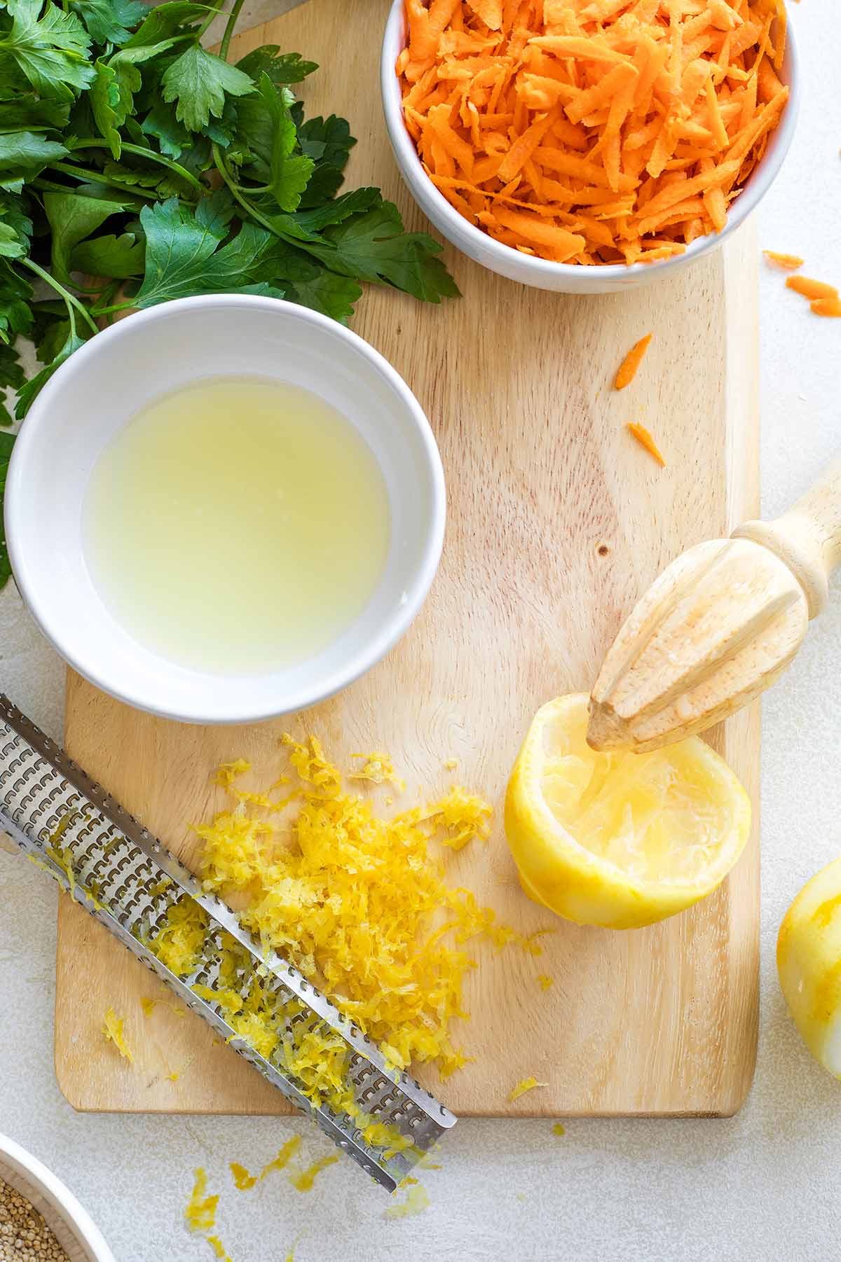 Half lemon on cutting board with zester and pile of zest, juicer and bowl of juice; plus additional ingredients nearby.