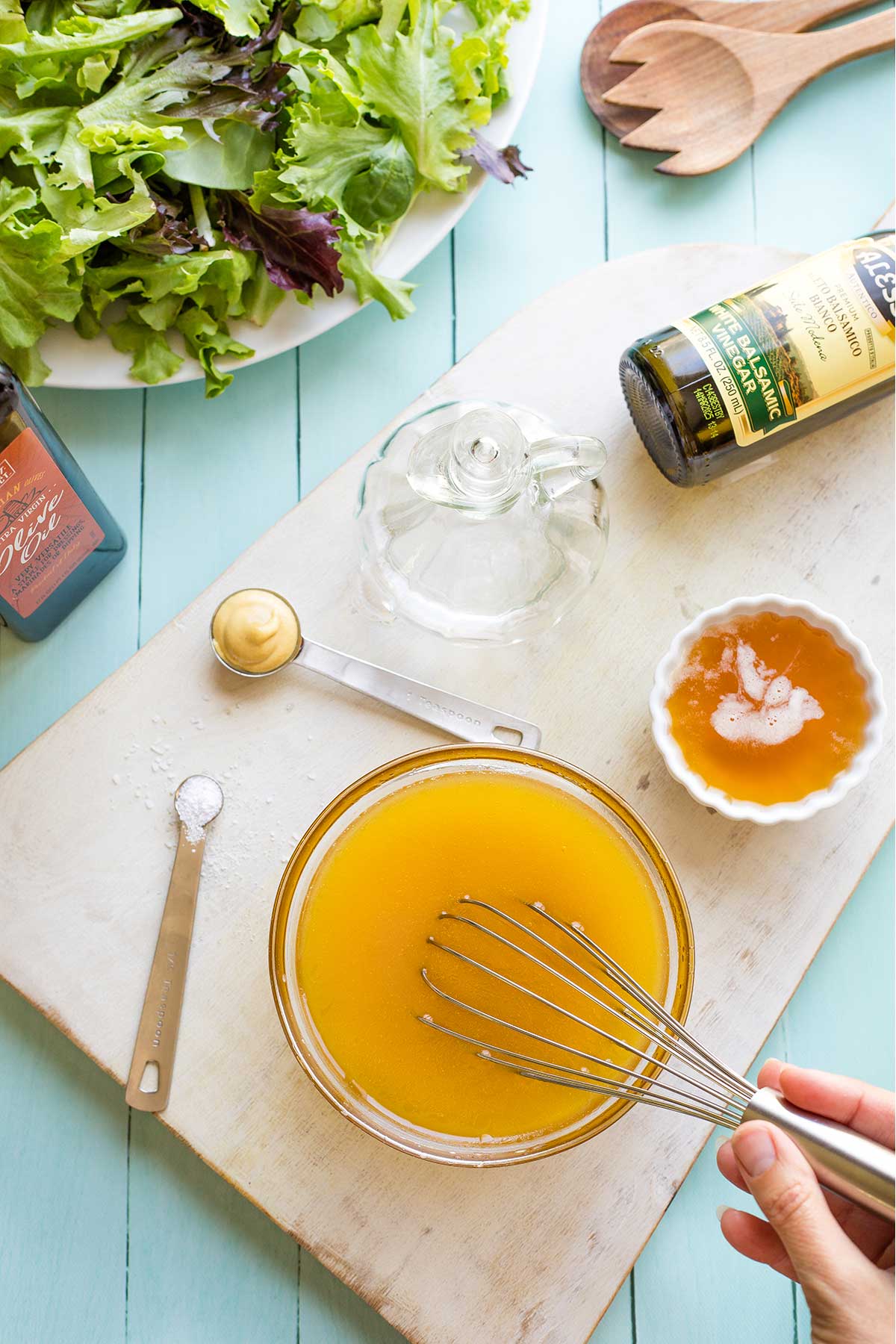 Overhead of a hand whisking together vinaigrette with other ingredients, and bottled olive oil and white balsamic vinegar surrounding.