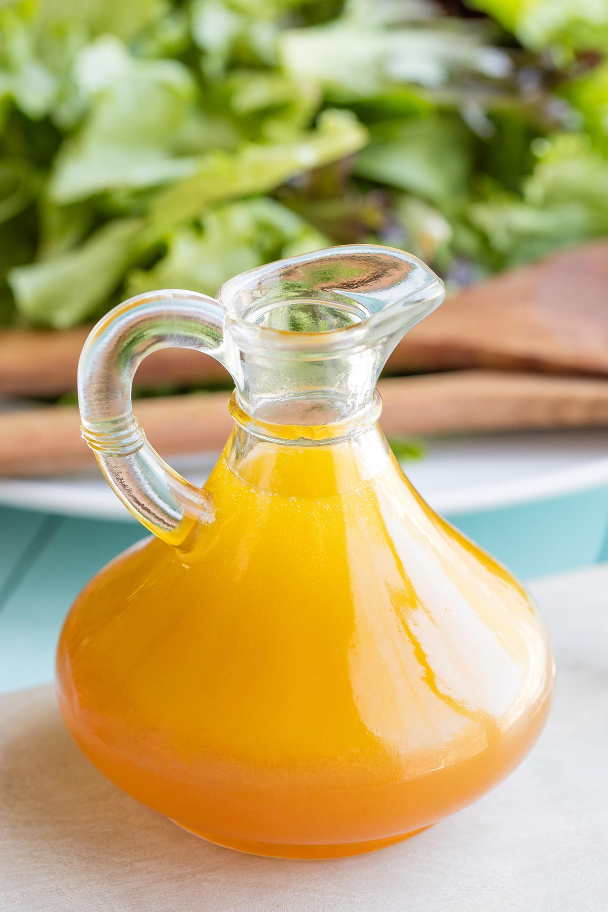 Side view of the vinaigrette in a small glass pitcher with salad greens in background.