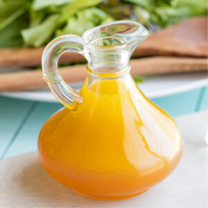 Vinaigrette in a cruet with wooden salad servers nestled into green salad behind.