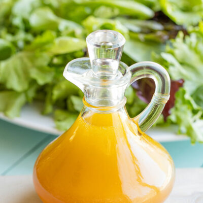 Side view of dressing in a clear, glass cruet with large plate of green salad in background.