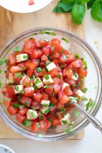 Overhead of glass bowl on cutting board, filled with Caprese topping with spoon.