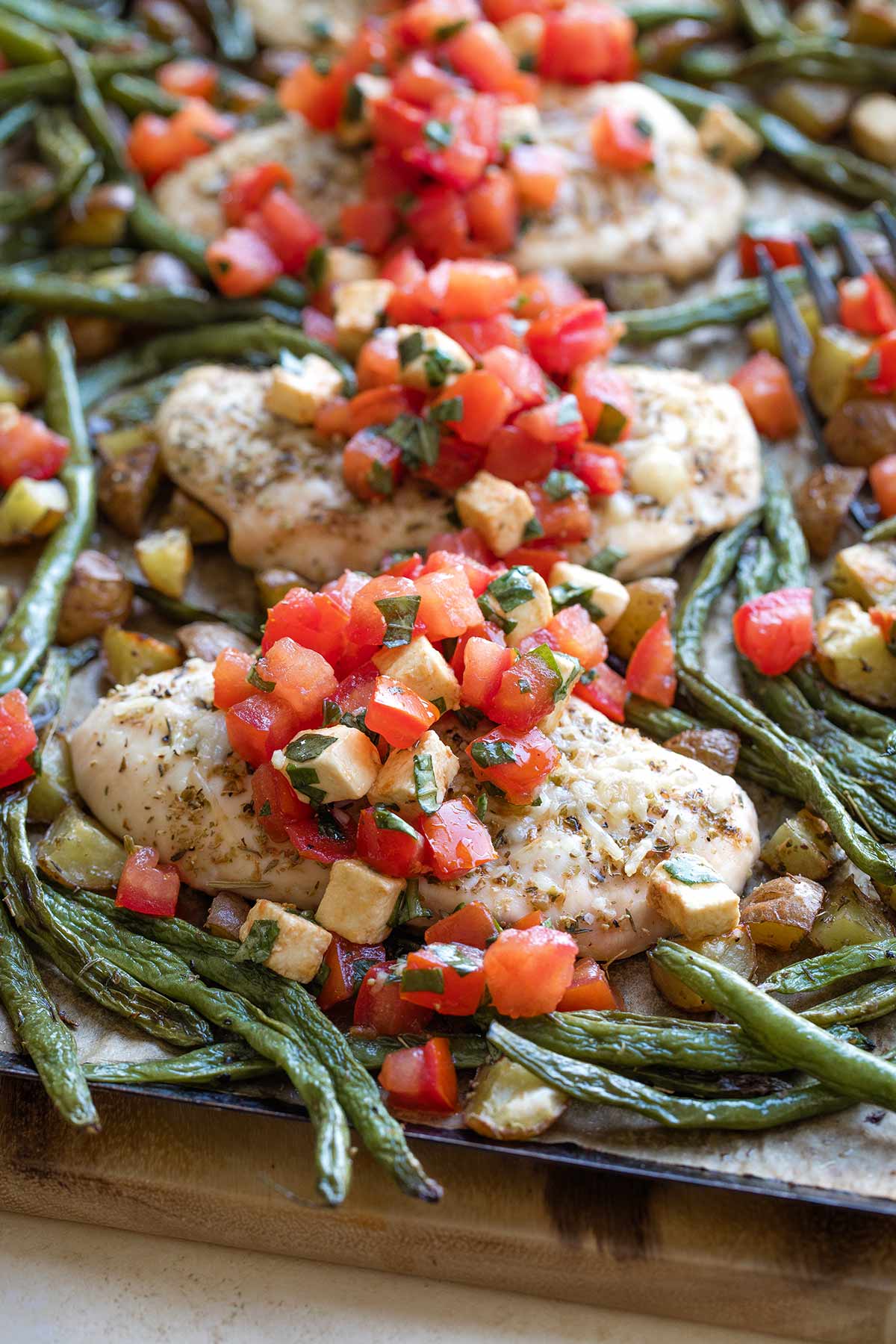 Side closeup of baked chicken breast surrounded by vegetables on pan with Caprese topping cascading across.