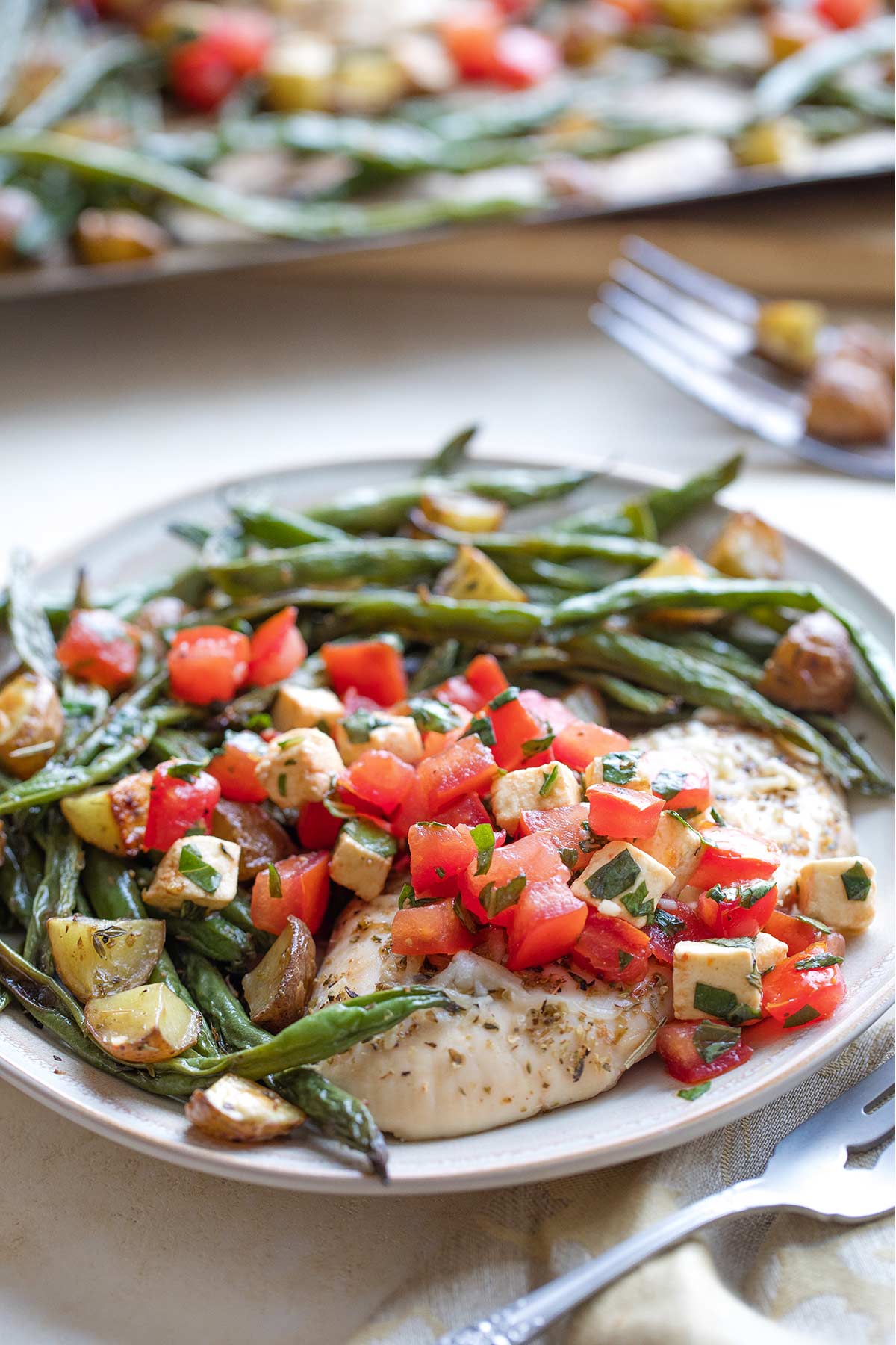 One plated dinner with baked chicken and vegetables with topping arranged on plate; pan with more in background.