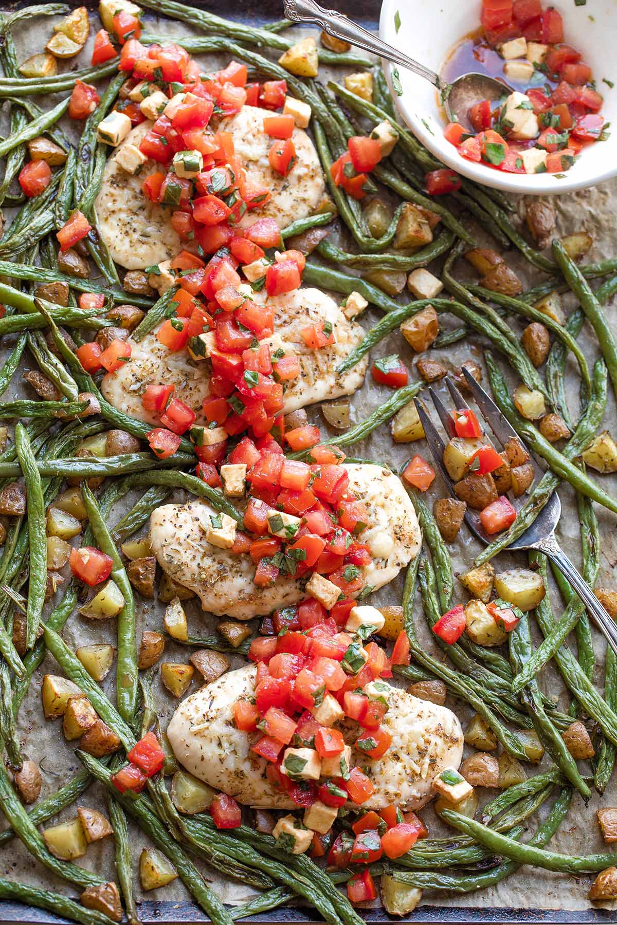 Flatlay of baked chicken and vegetables combined on 1 sheet pan with topping and serving fork.