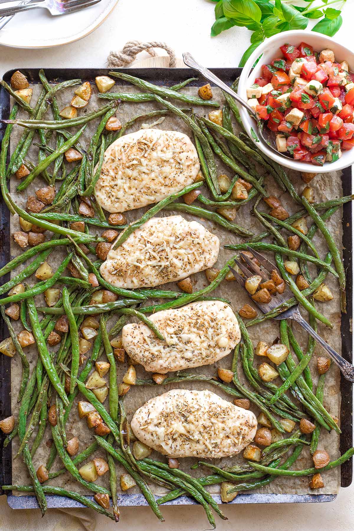 Overhead of baked chicken and vegetables combined onto one sheet pan; Caprese topping in bowl ready to be drizzled on top.