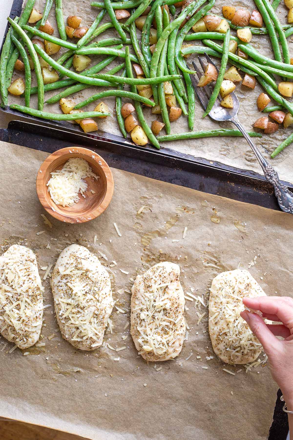 Hand sprinkling parmesan cheese on partially baked chicken; sheet pan of stirred vegetables behind.