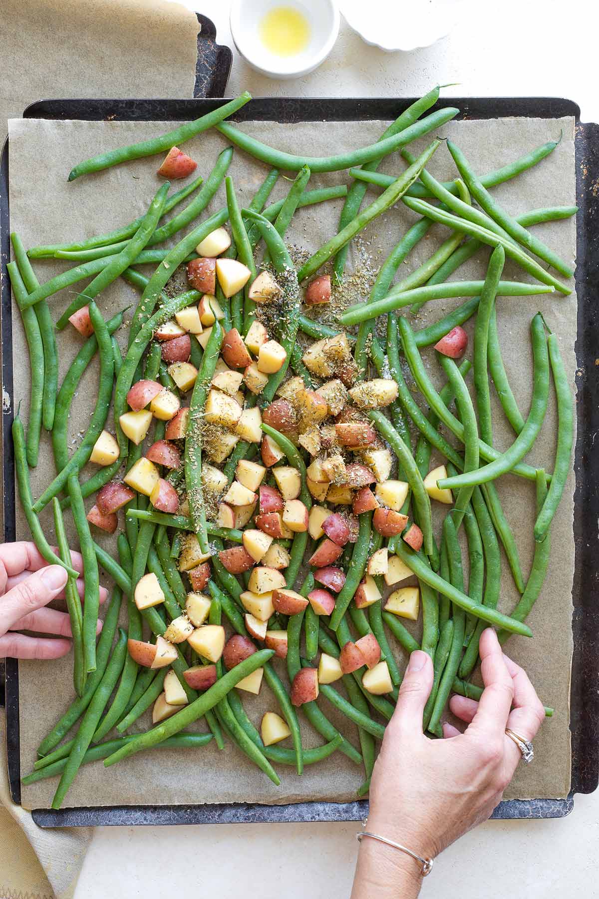 Two hands tossing the vegetables with seasonings and olive oil on parchment-lined sheet pan.