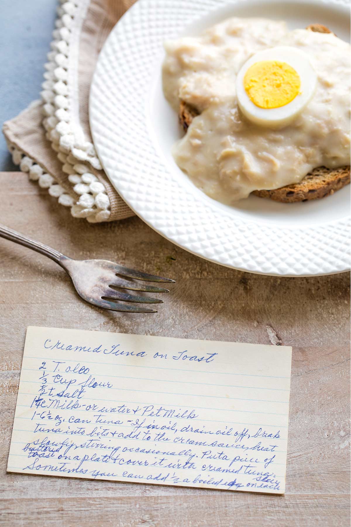 My Grandma's Creamed Tuna on Toast recipe card on a cutting board in front of her variation of serving it with a hard-boiled egg.