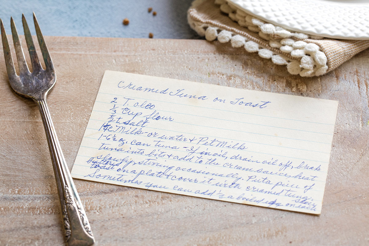 My Grandma's hand-written recipe card for Creamed Tuna on Toast, on a cutting board with fork alongside.