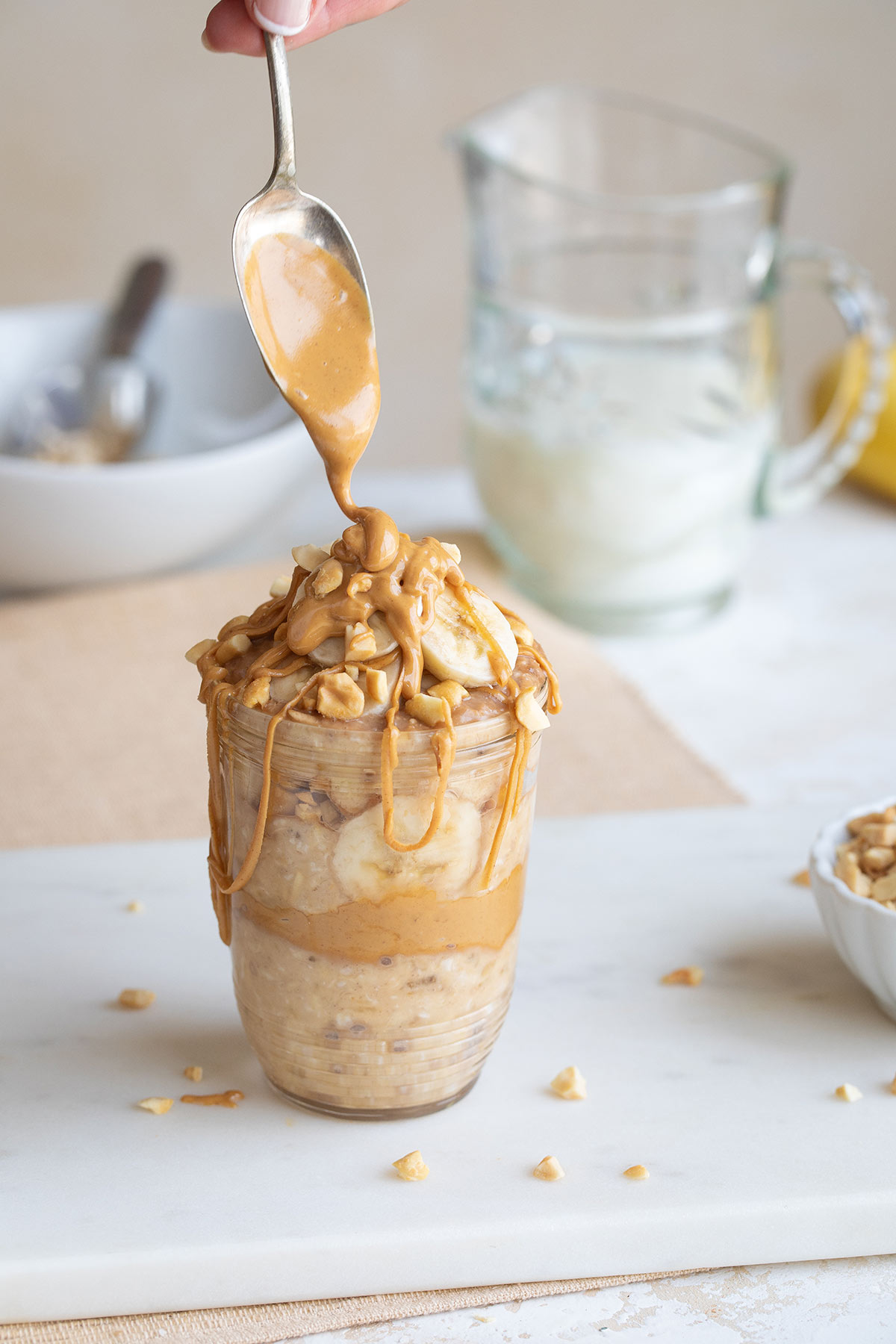 Hand holding spoon above jar of oats to drizzle warmed peanut butter on top.