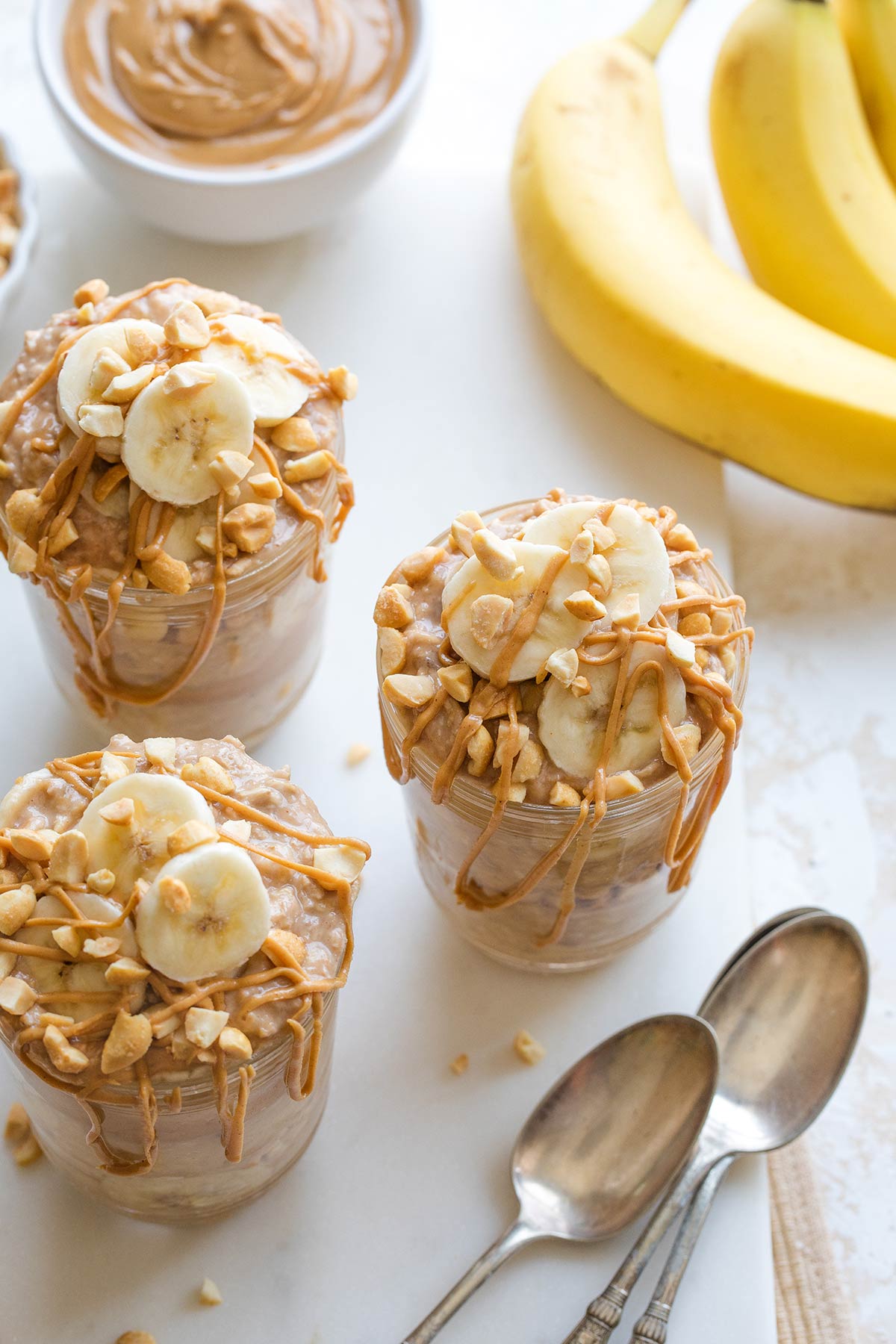 Top down of three servings with spoons, whole unpeeled bananas and small bowl of peanut butter surrounding.