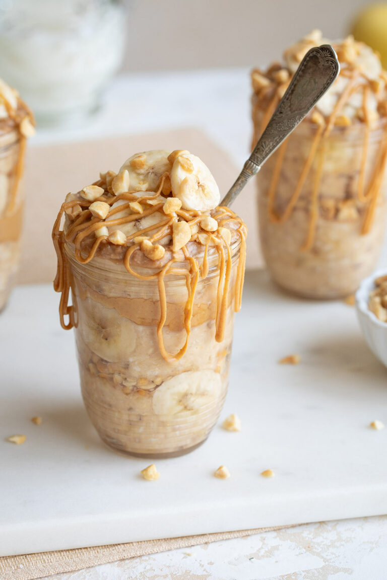 Side closeup of one serving in pretty jar, garnished with sliced bananas and chopped nuts, with peanut butter drizzle and spoon jutting out.