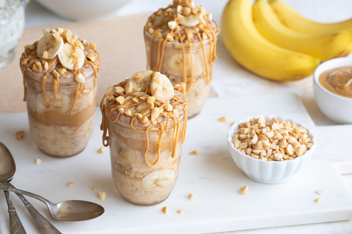 3 servings in glass jars surrounded by spoons, bananas, bowls of peanut butter and nuts.