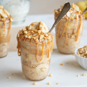 Side closeup of one jar of overnight oats, topped with nuts and bananas, drizzled with peanut butter, with spoon tucked in.