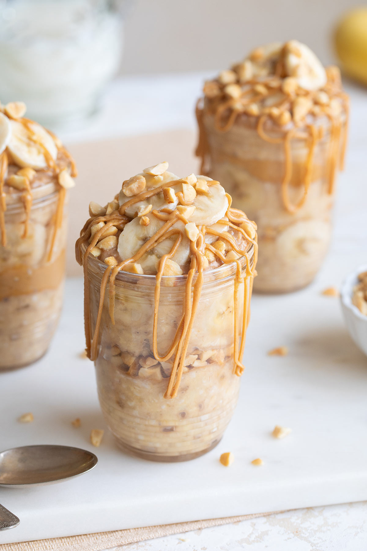 Three glass mason jars on white cutting board, full of oats that have been topped with sliced bananas and drizzled with peanut butter.