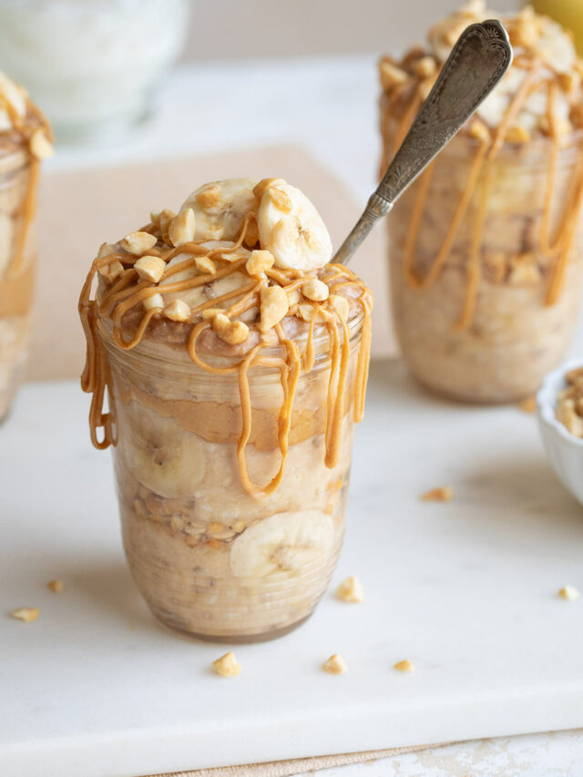 Side closeup of one serving in pretty jar, garnished with sliced bananas and chopped nuts, with peanut butter drizzle and spoon jutting out.