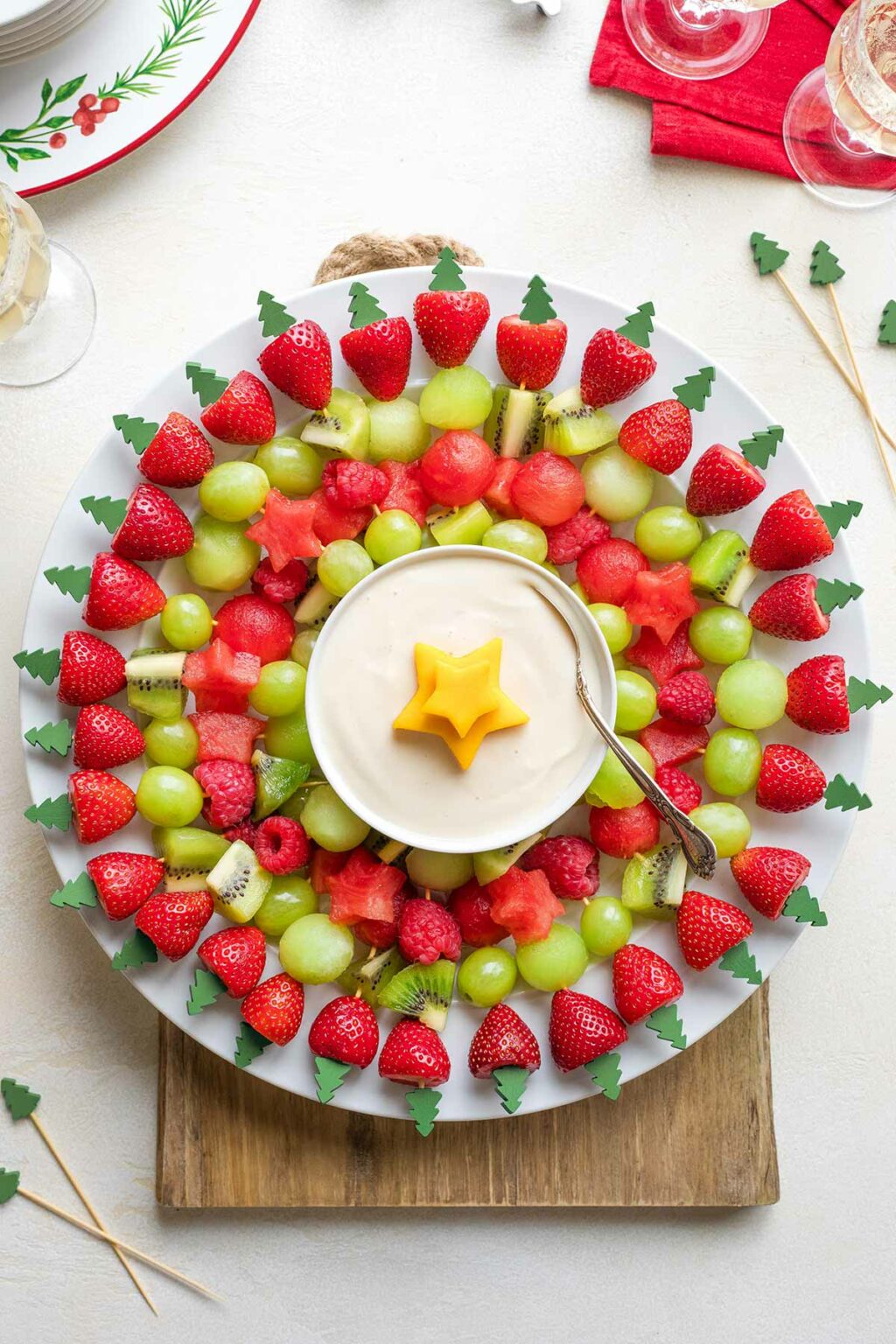 Flatlay of wreath on white plate on wooden board with extra kabobs, Christmas plates and napkin and wine glasses nearby.