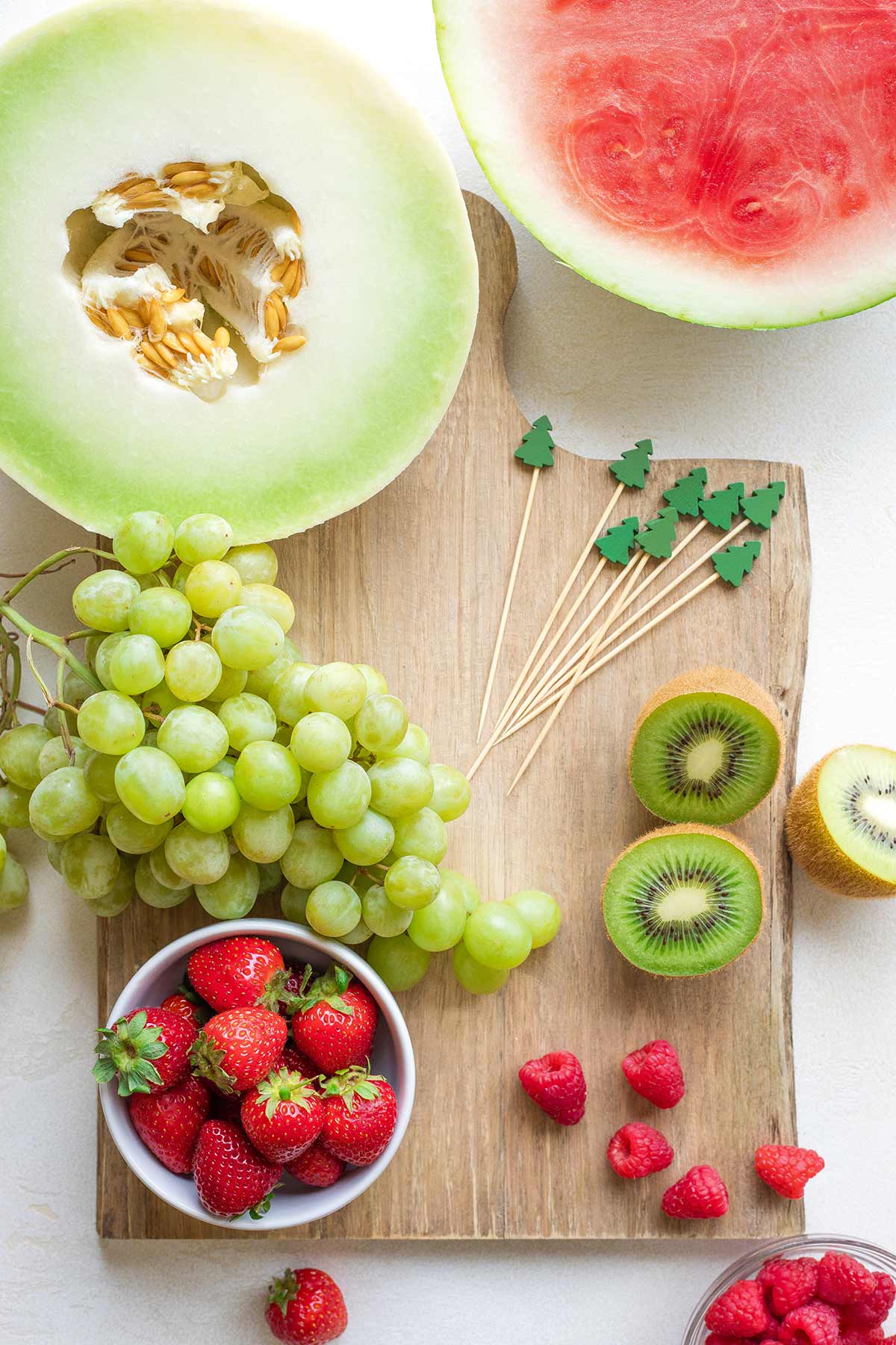 Flatlay of various red and green fruits on cutting board: halved melons, a bunch of grapes, halved kiwi, red berries.