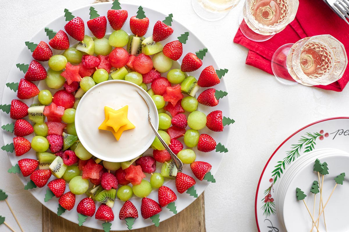 Christmas fruit wreath toward left side, with stack of party plates, extra kabob skewers, red napkin and glasses of wine at right.