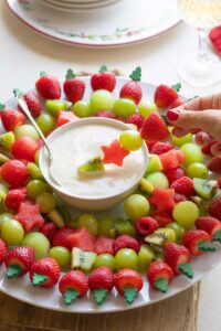 Party scene of fingers dipping a skewer from wreath into central dip bowl; Christmas plate, red napkin, and glass of wine in background.