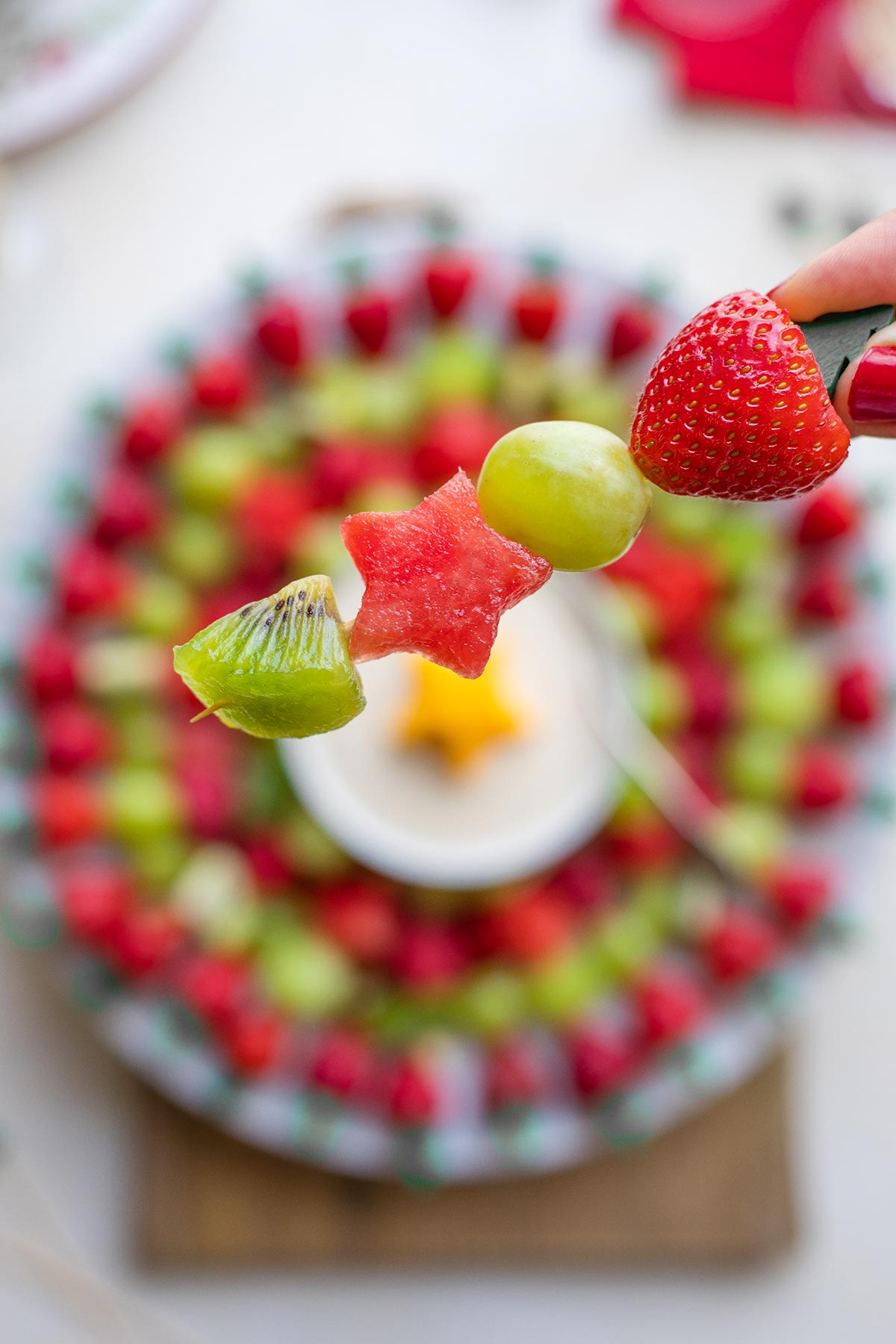 Closeup of fingers holding one fruit skewer with a kiwi chunk, star-shaped watermelon, green grape and strawberry.