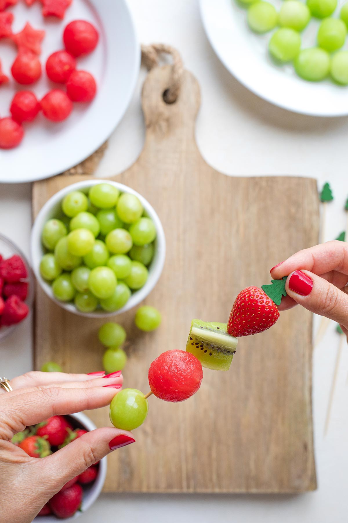 Two hands threading fruit onto a kabob skewer with other fruits visible in background beneath.