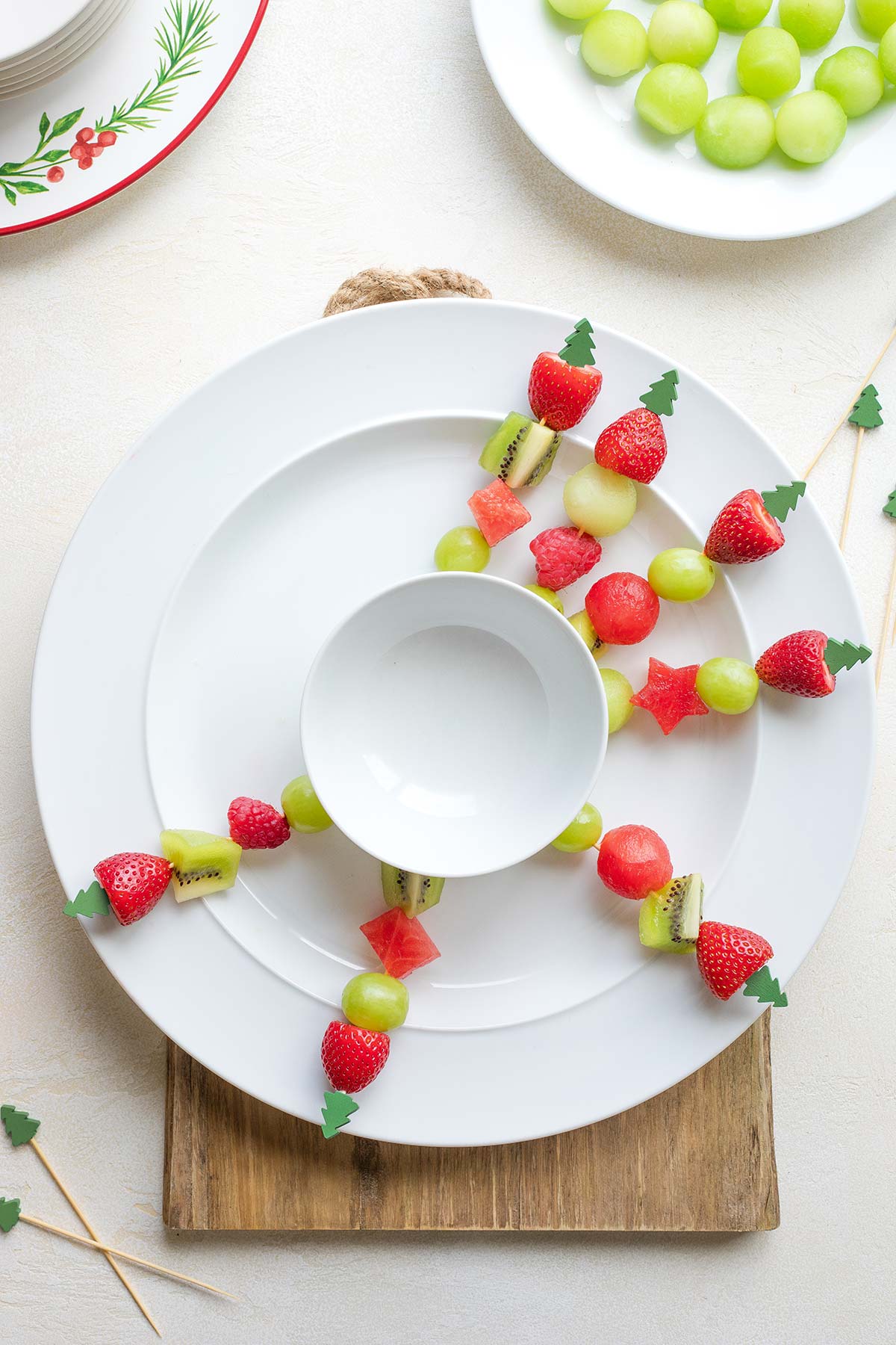 Starting to build wreath - 7 kabobs topped with green Christmas trees are laid on white plate around empty white bowl at center.