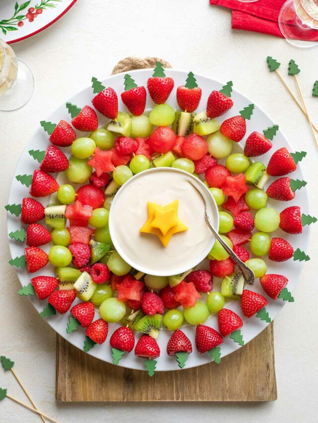 Flatlay of wreath on white plate on wooden board with extra kabobs, Christmas plates and napkin and wine glasses nearby.