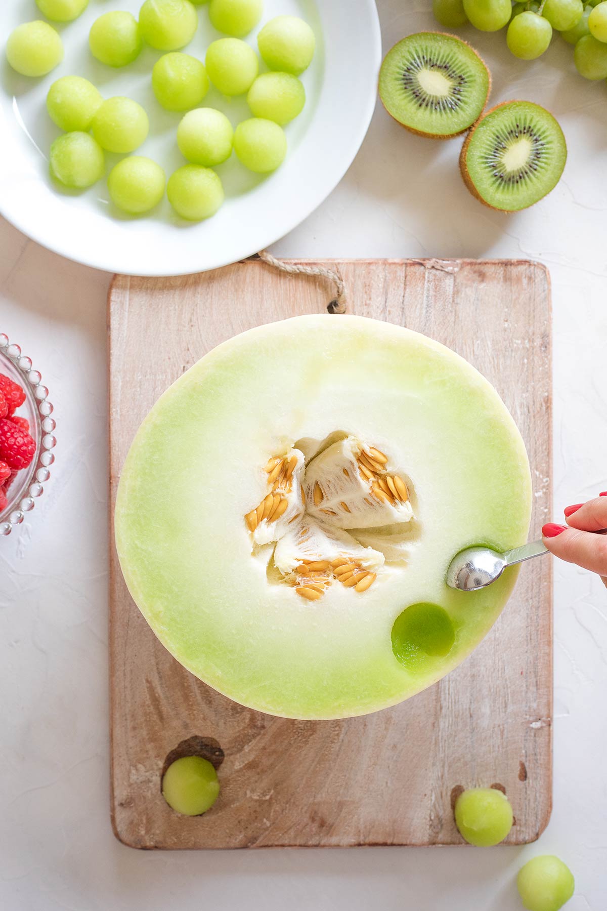 Hand using measuring spoon to shape melon balls out of half a honeydew.