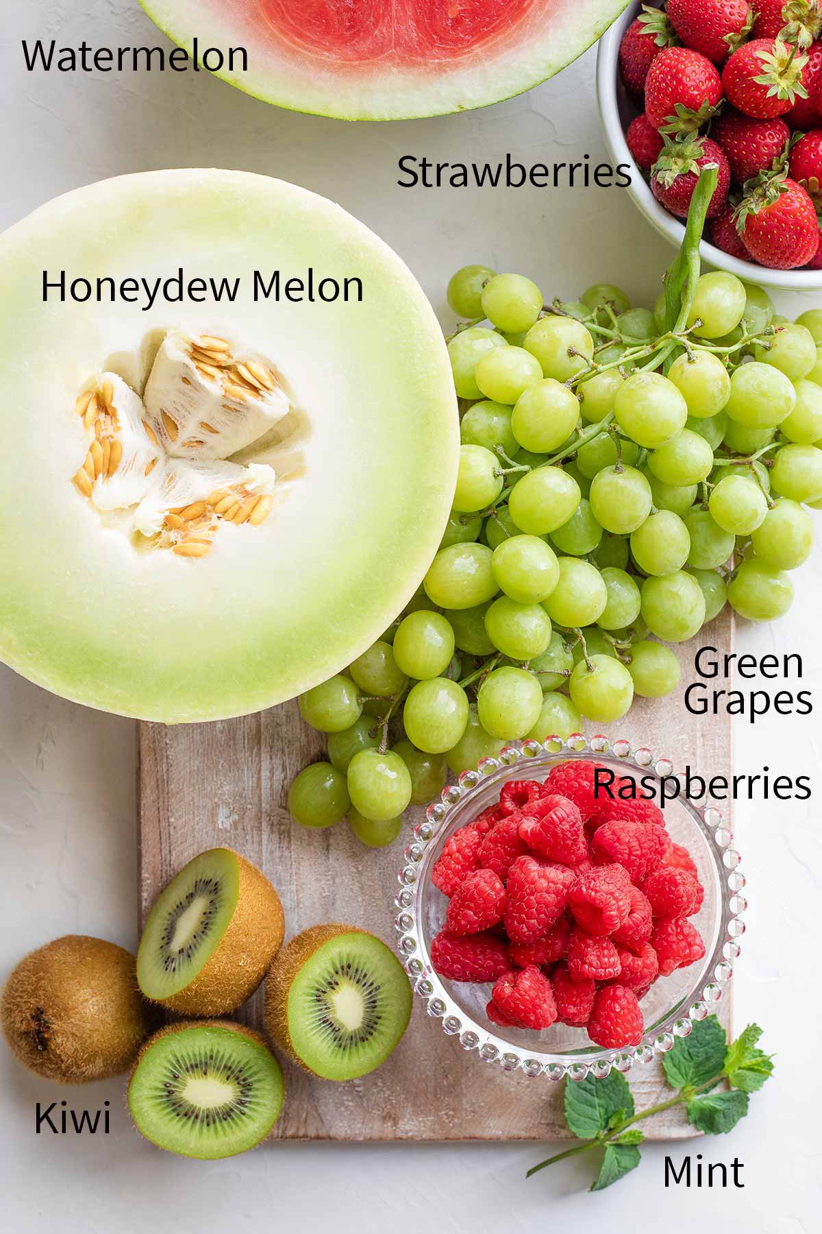 Ingredients arranged on cutting board and labeled (clockwise) Watermelon, Strawberries, Green Grapes, Raspberries, Mint, Kiwi, Honeydew Melon.