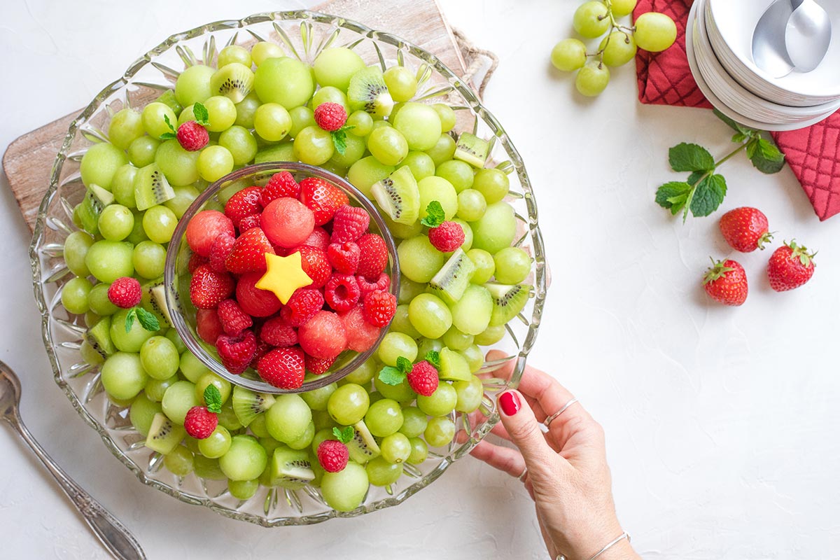 One hand holding the fruit platter; additional fruit and Christmas napkin, bowls and spoons at corner.