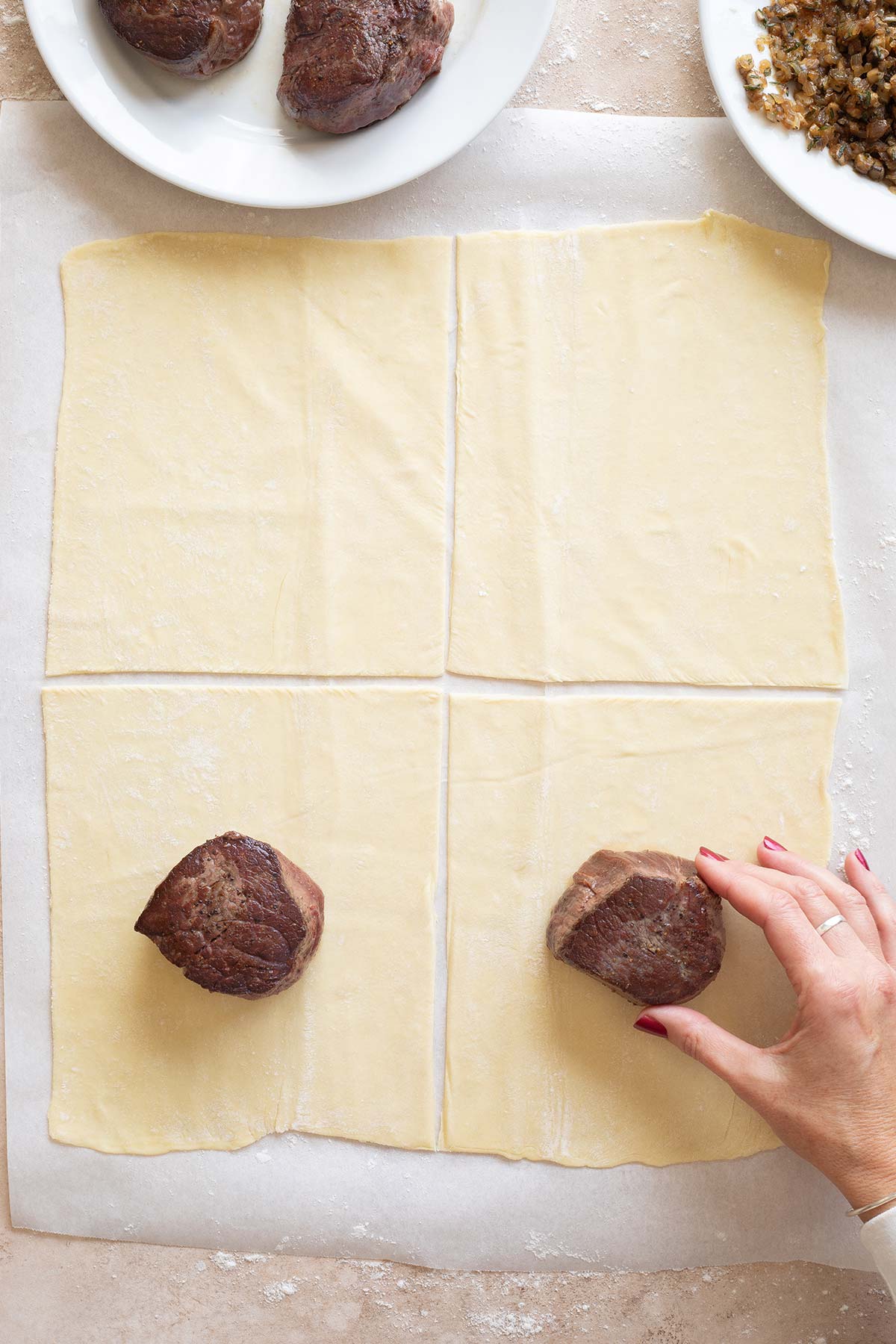 Hand placing a seared beef filet mignon in the center of each puff pastry section.