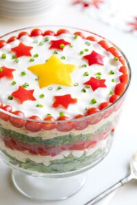 Salad presented in glass trifle bowl so you can see the 7 layers; serving spoon, potluck plates and Christmas decoration nearby.