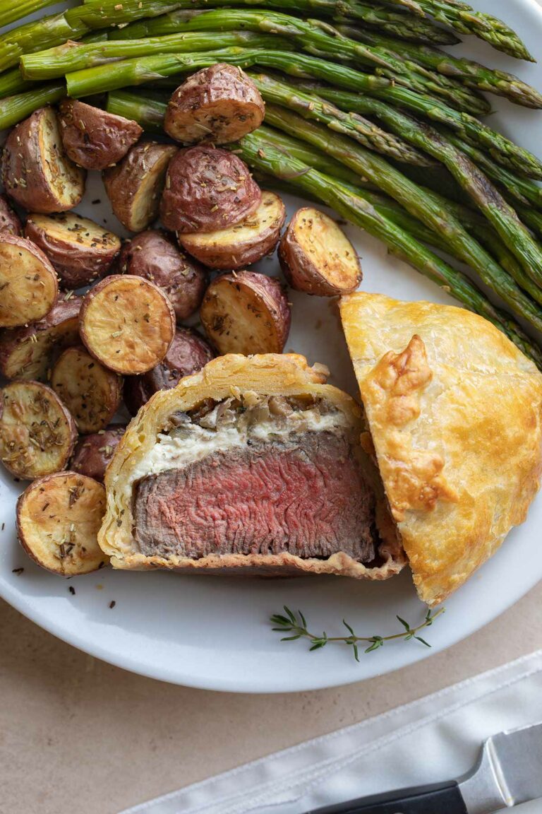 Flatlay of an individual beef Wellington, cut in half with one half facing upwards, surrounded on white plate by roasted asparagus and potatoes.