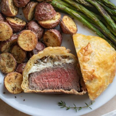 Overhead of one cut Wellington with half tipped up so you can see inner filling, on plate with asparagus and potatoes.