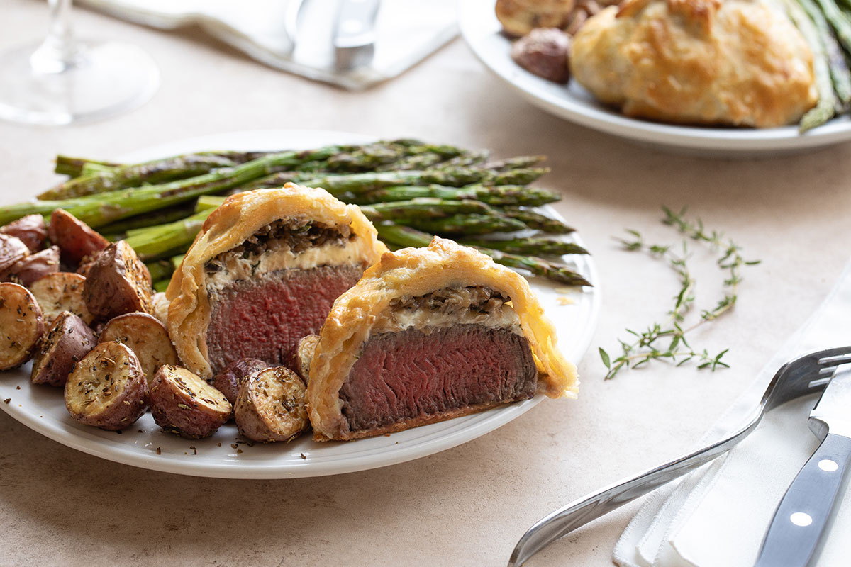 Dinner scene of beef Wellington recipe for two people, each plated with asparagus and red potatoes, with silverware and wine glass nearby.