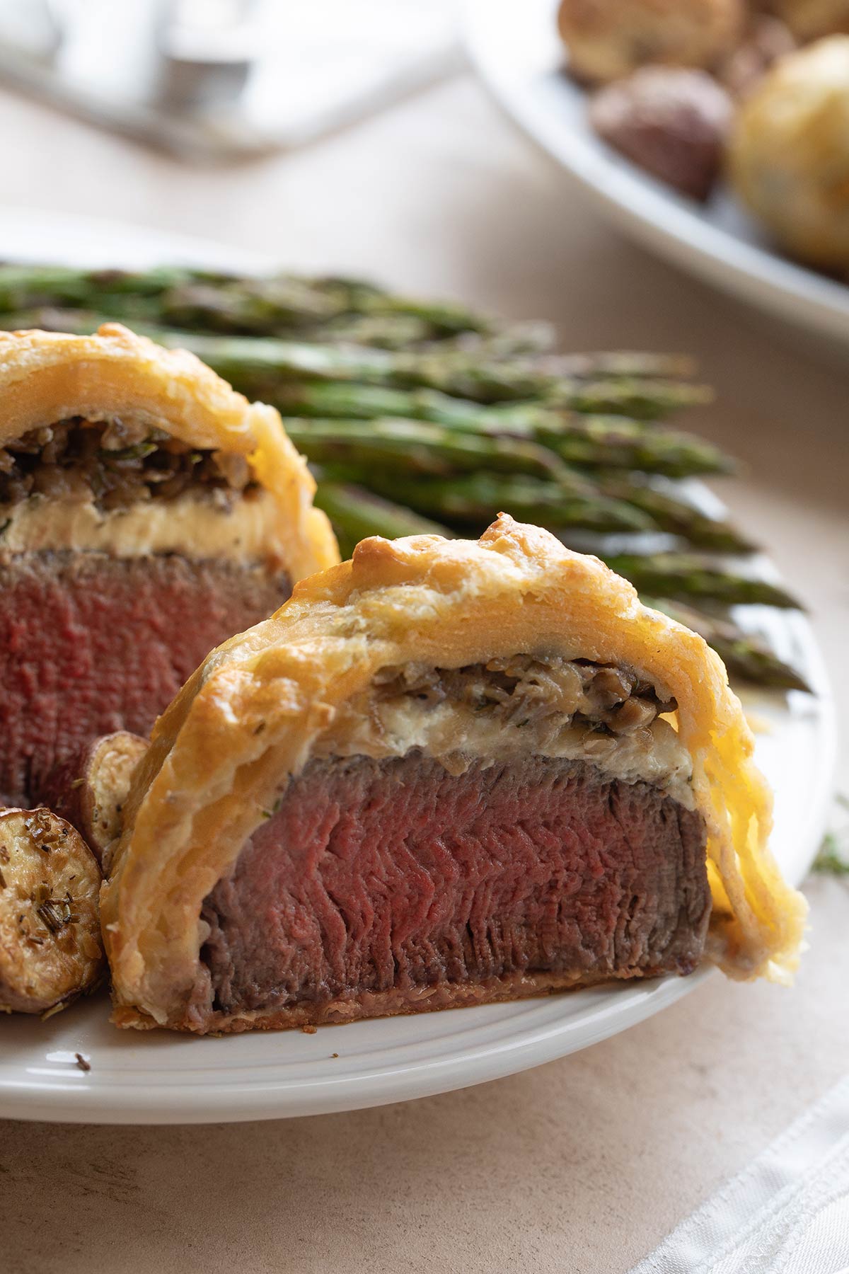 Closeup of half of a mini Wellington at edge of dinner plate, surrounded by vegetables with second plate in background.