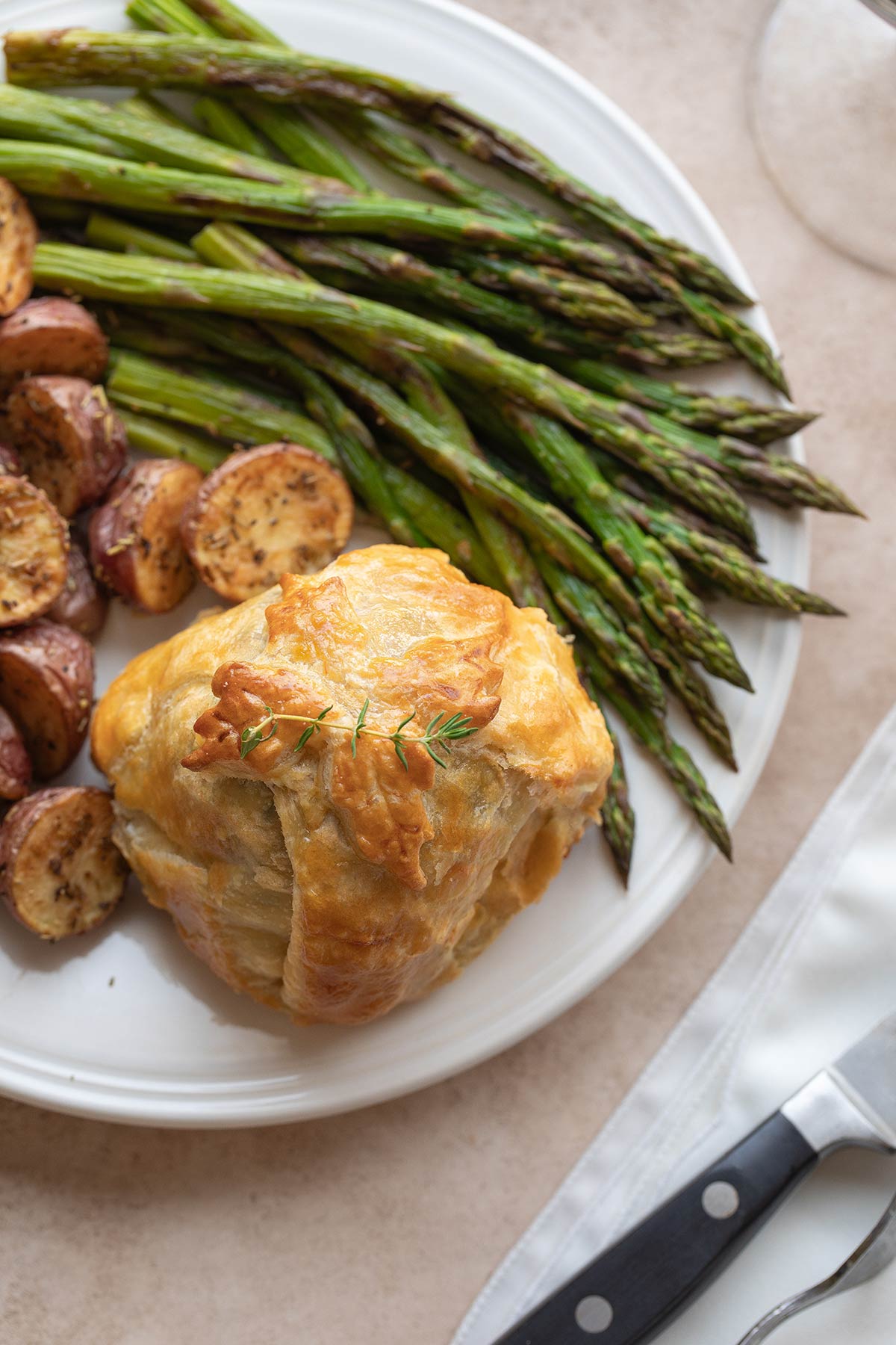 Flatlay of an individual beef Wellington, served uncut with vegetables and decorative thyme sprig.