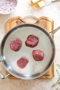 Overhead of 4 raw filet mignon steaks, salted and peppered, added to skillet to sear.