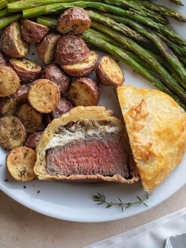 Flatlay of an individual beef Wellington, cut in half with one half facing upwards, surrounded on white plate by roasted asparagus and potatoes.